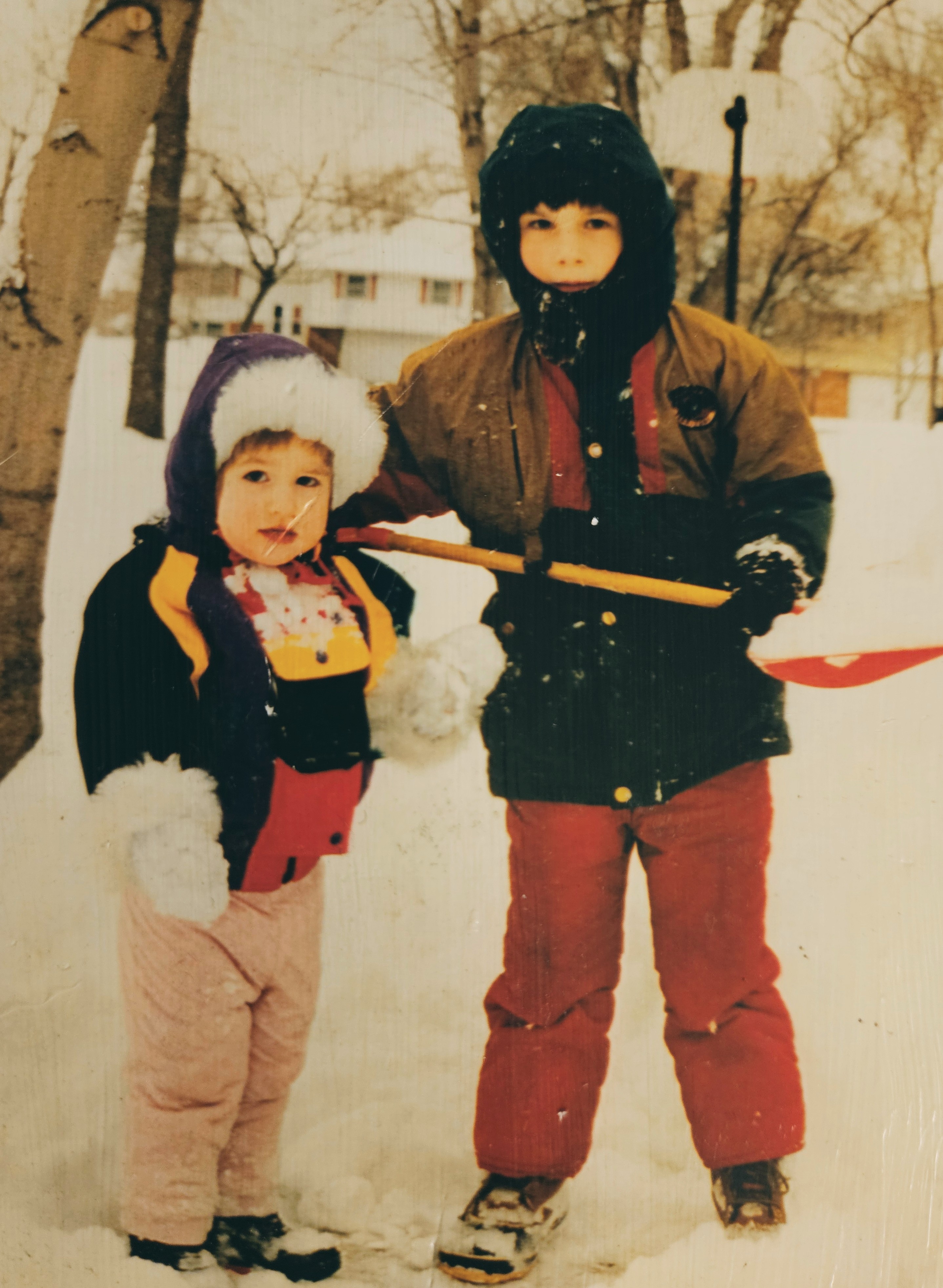 Two small children are very bundled up in winter gear, one of them is holding a shovel, they are surrounded by a snowy suburbia